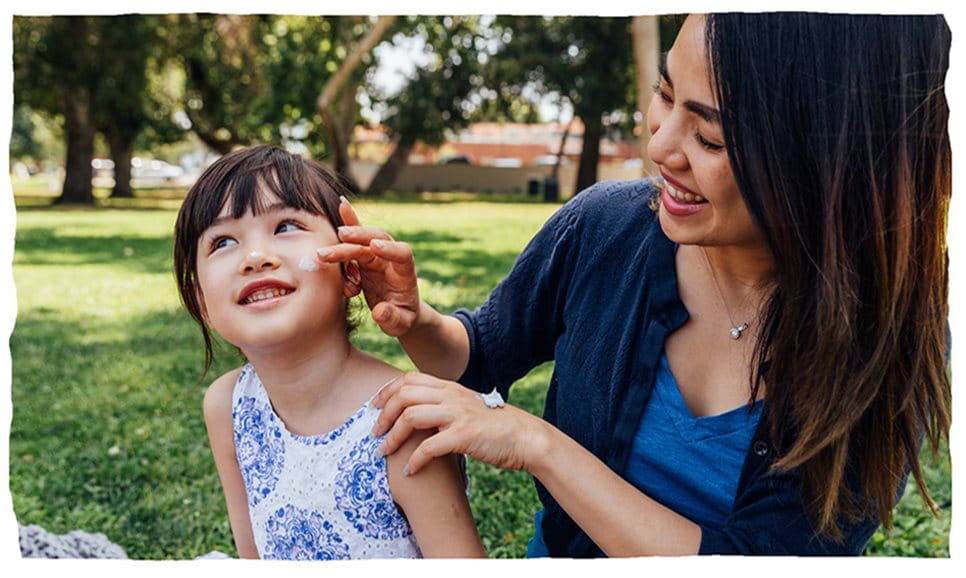 En kvinna applicerar lotion på ett barns ansikte i en park.