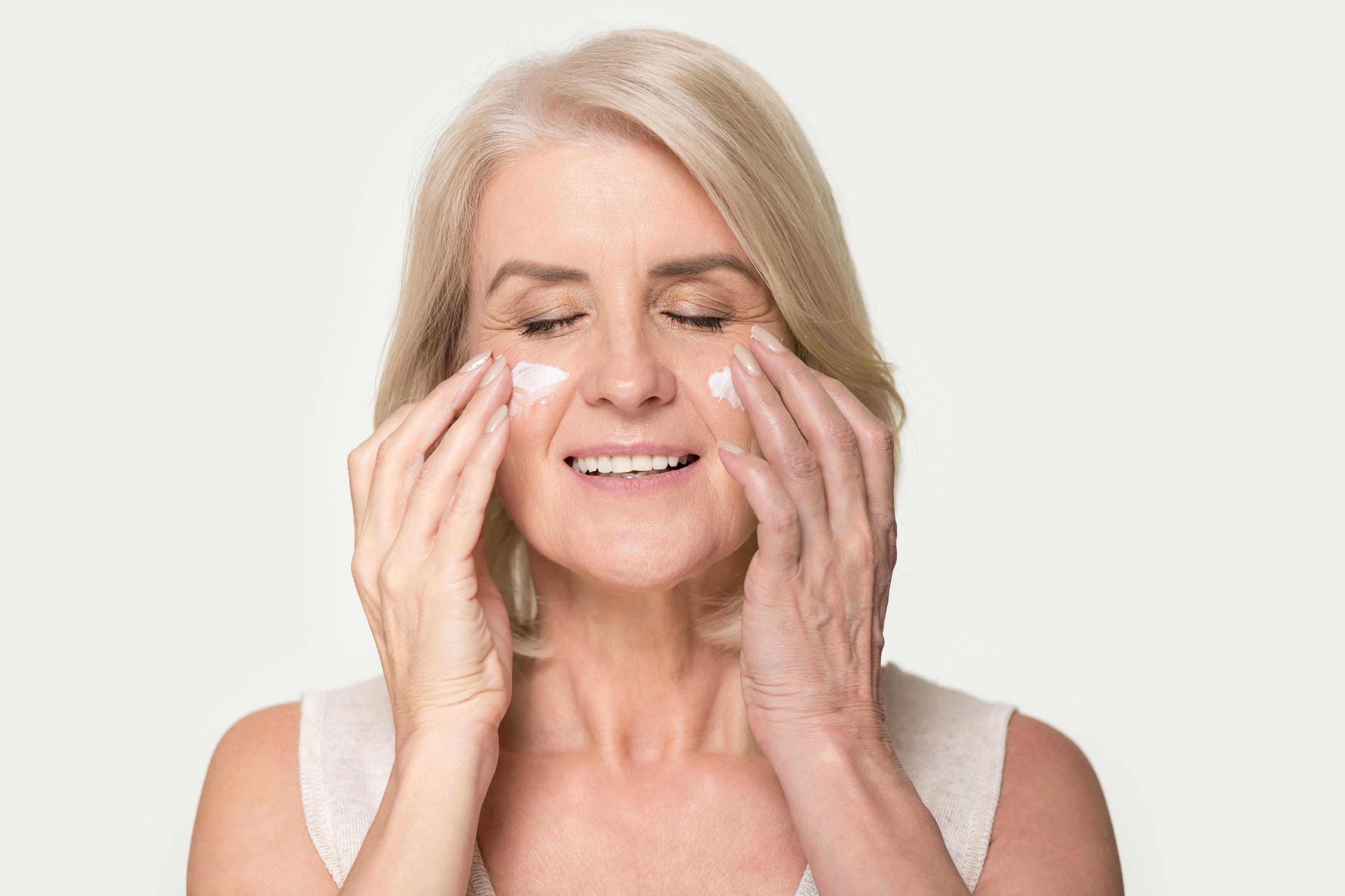 Eine ältere Frau cremt ihre Wangen mit einer Creme ein und lächelt mit geschlossenen Augen.