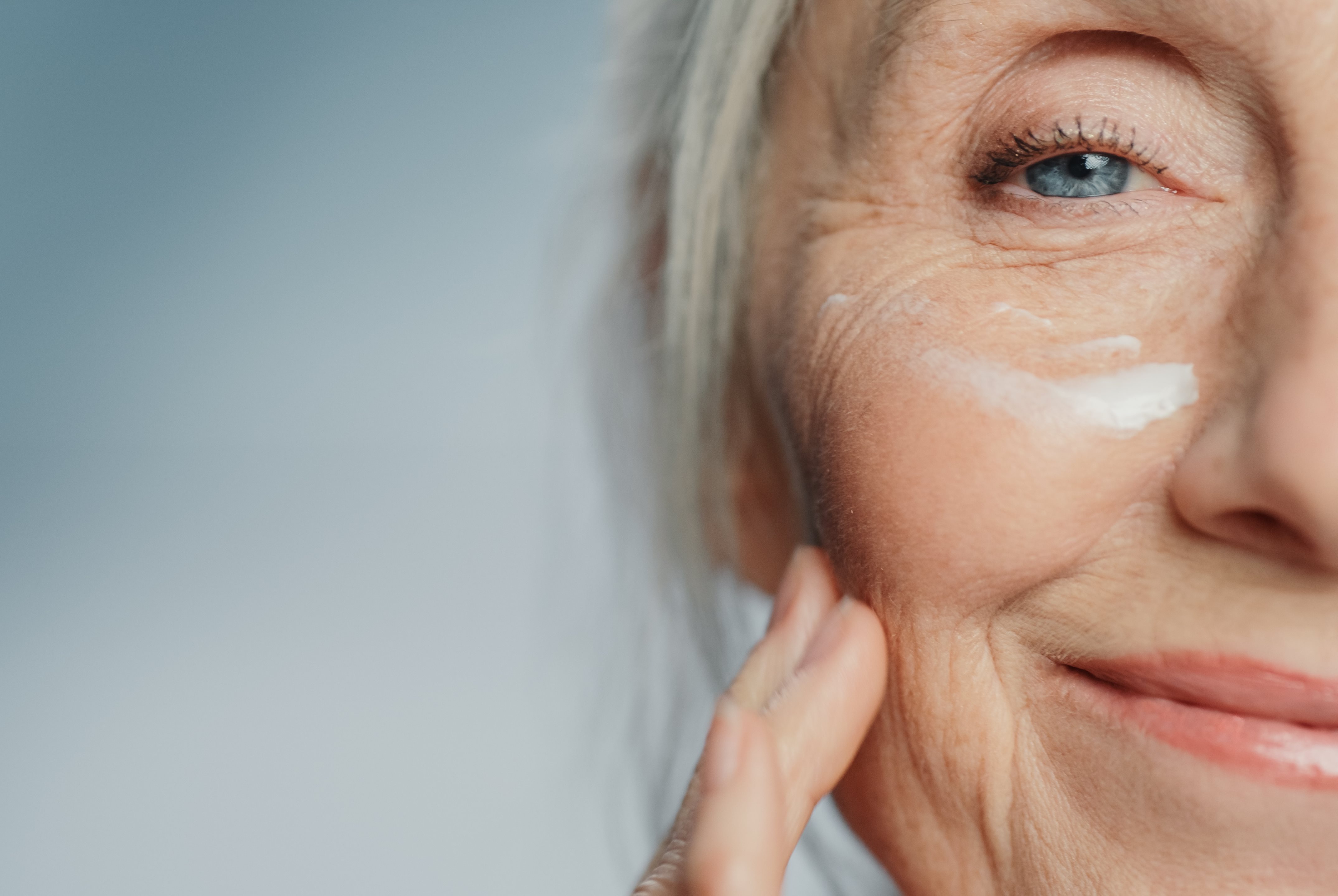 Nahaufnahme der Gesichtshälfte einer lächelnden älteren Frau mit sichtbaren Falten, die sich Creme auf die Wange aufträgt.