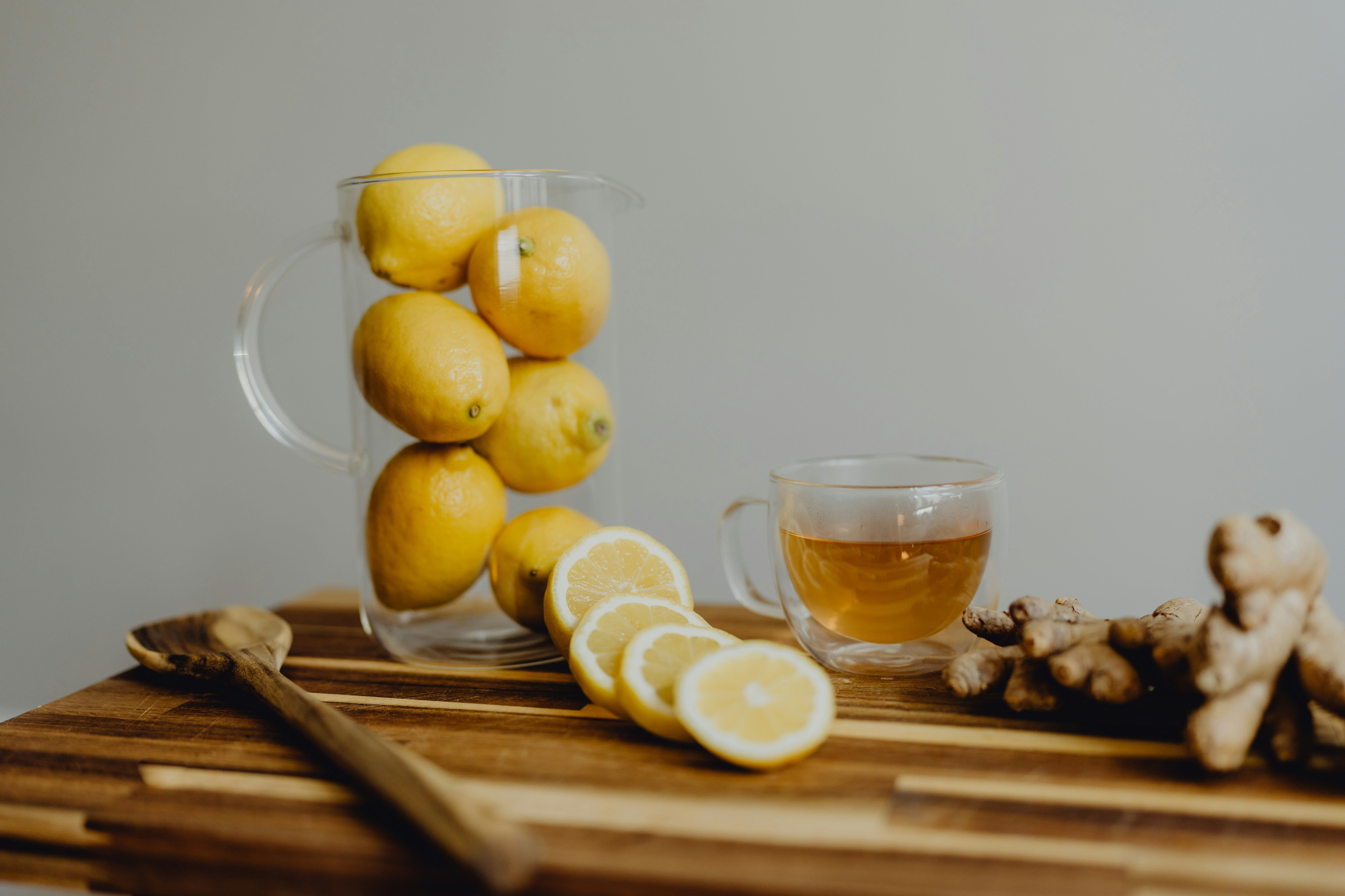 Eine Holztischplatte, auf der sich ein Glas mit mehreren Zitronen, einige aufgeschnittene Zitronen, ein Glas mit einer hellbraunen Flüssigkeit und einer Ingwerknolle befinden.
