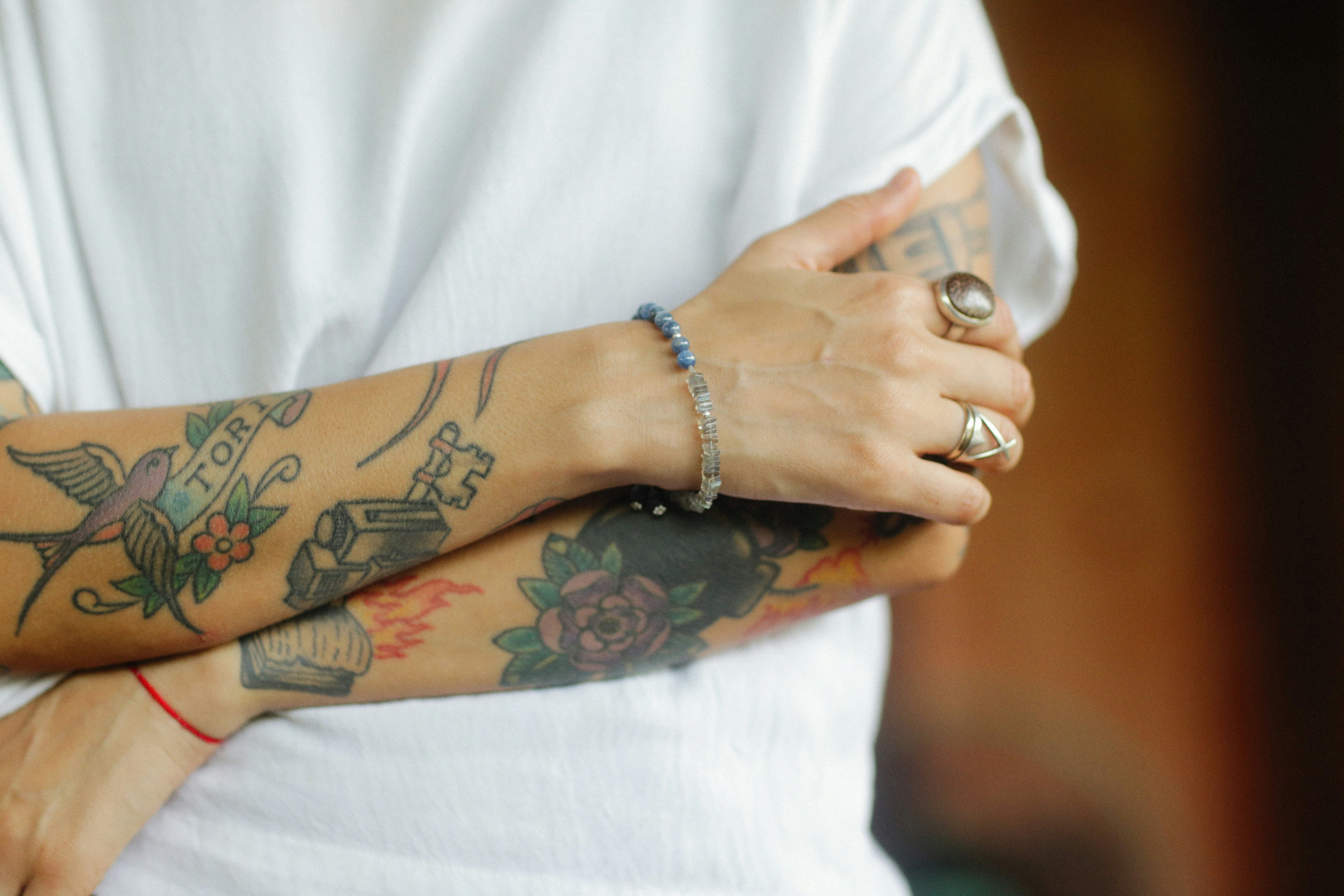 Person im weißen T-Shirt verschränkt die Arme. Ihre Unterarme sind mit Tattoos bedeckt und sie trägt Schmuck an den Fingern und um das Handgelenk.v