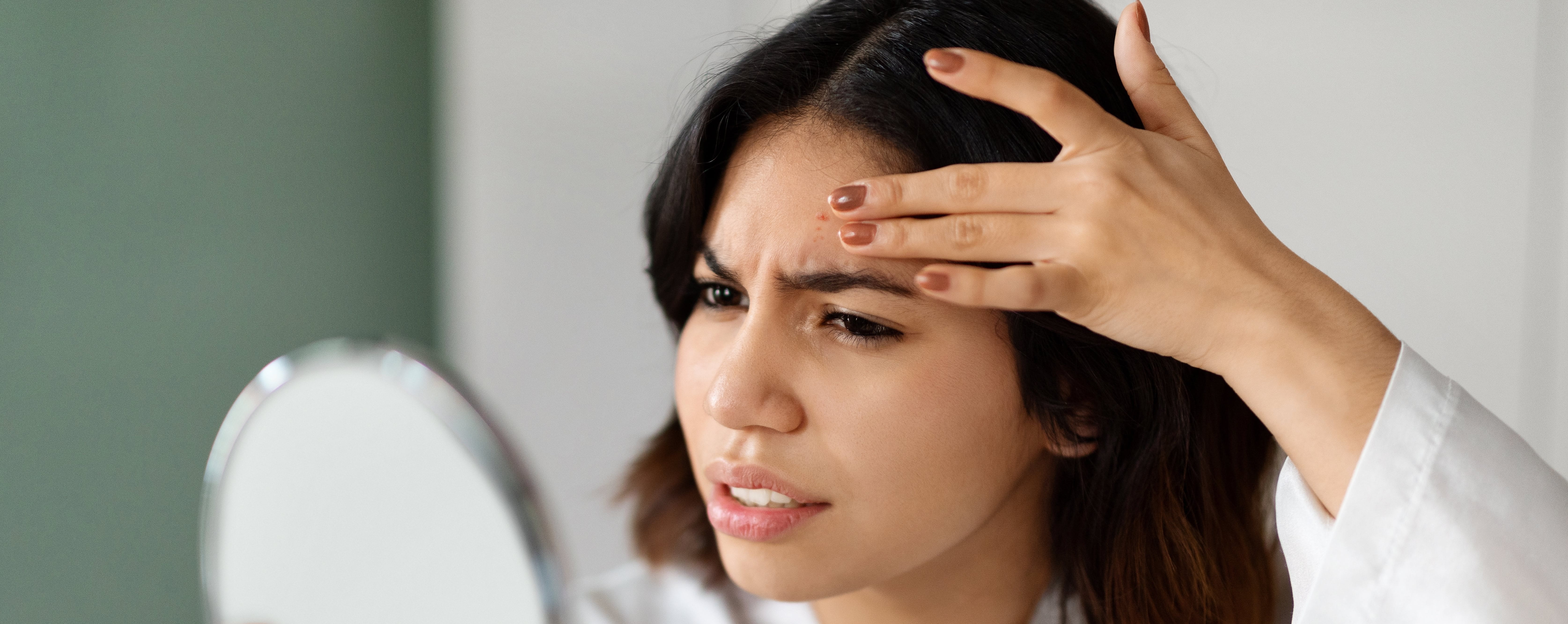 Frau schaut entsetzt in einen Handspiegel und fasst mit der Hand an einen Pickel auf die Stirn.