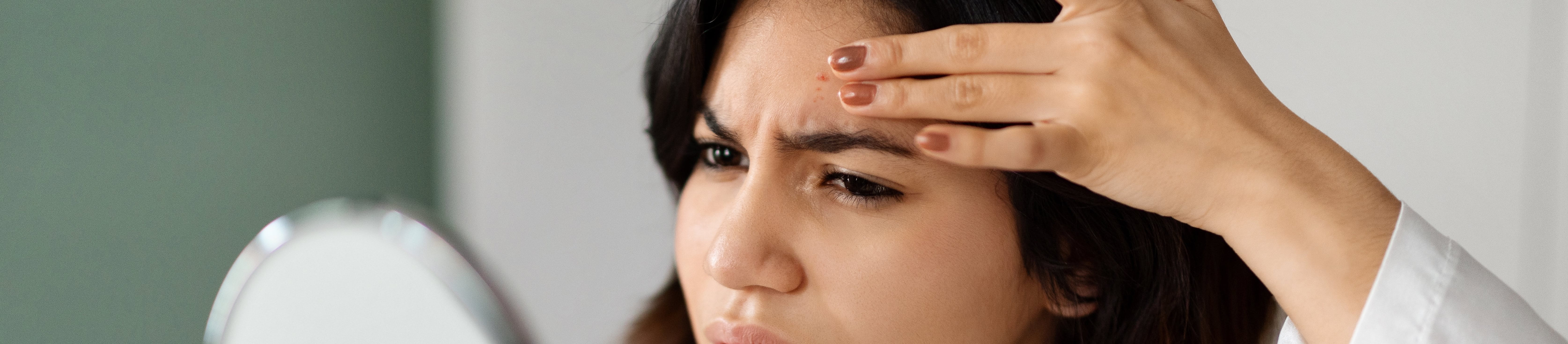 Frau schaut entsetzt in einen Handspiegel und fasst mit der Hand an einen Pickel auf die Stirn.