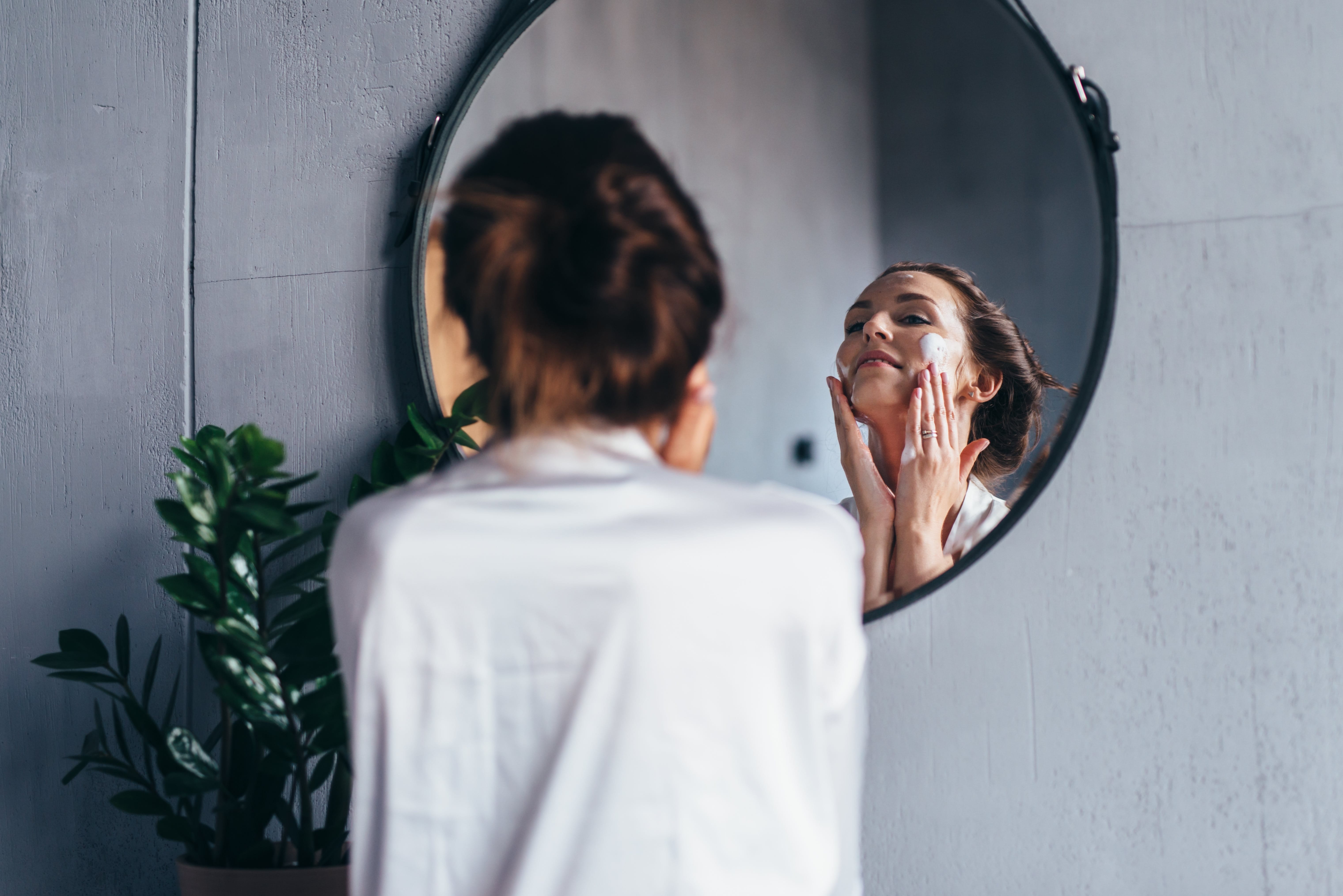 Frau blickt in einen runden Spiegel an einer grauen Wand. Sie trägt eine weiße Bluse, die braunen Haare zu einem Zopf gebunden und hat den Kopf in den Nacken gelegt. Mit den beiden Händen trägt sie Creme auf ihre Wangen auf.