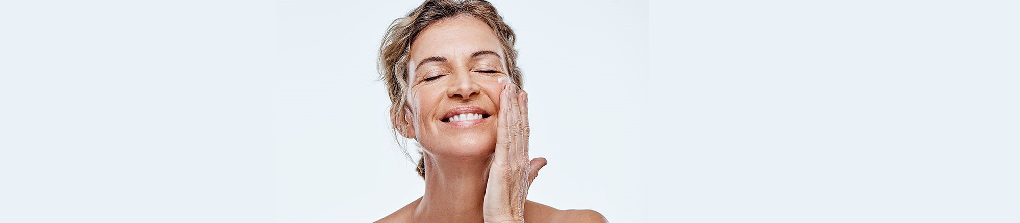 Donna matura sorride mentre applica una crema contorno occhi