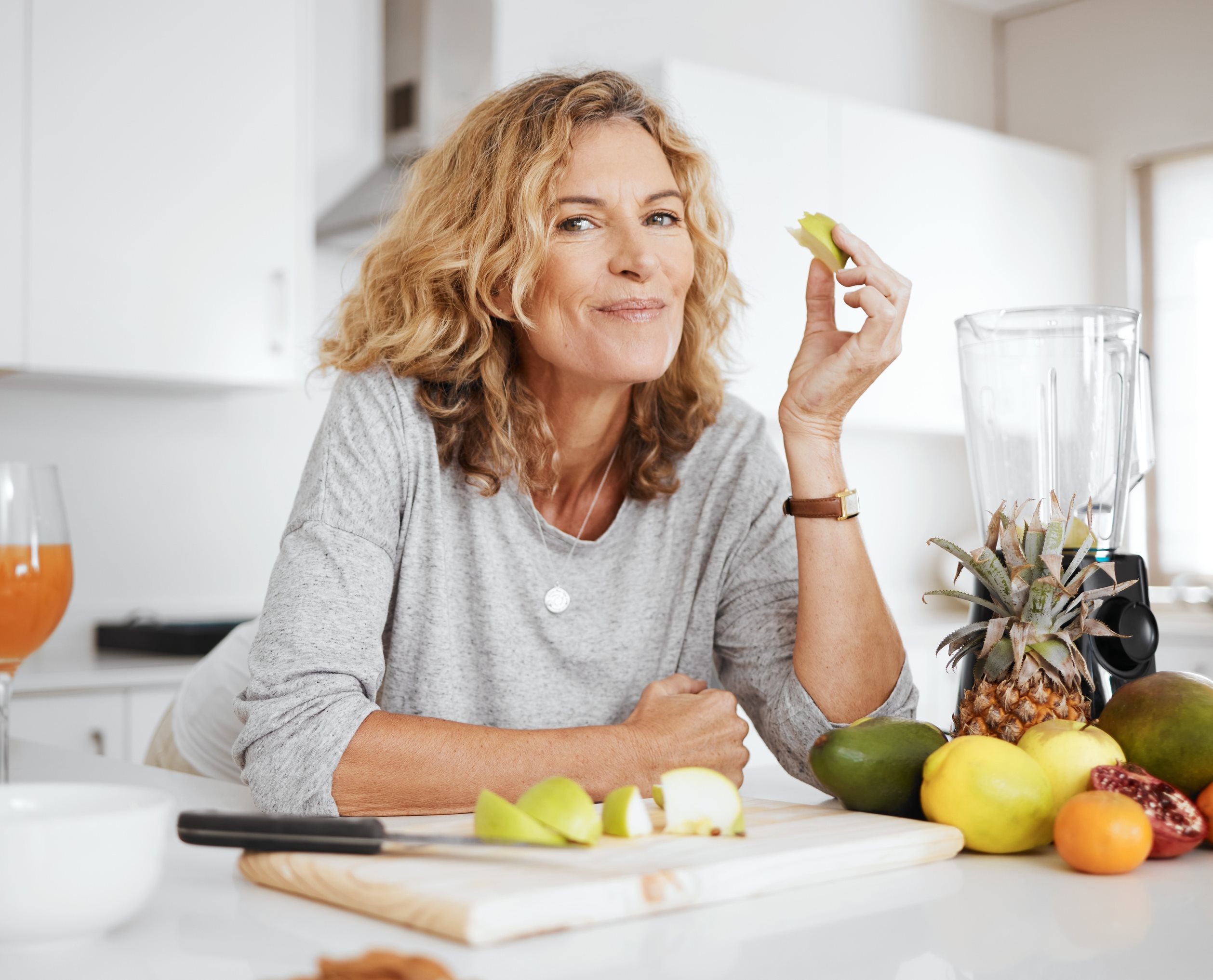 Eine Frau isst ein Stück Apfel. Auf dem Küchentresen vor ihr liegt noch weiteres Obst.