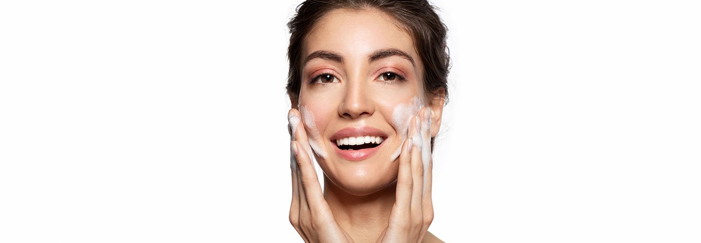 Ragazza sorride mentre massaggia il viso con un detergente schiumoso durante la doppia detersione quotidiana.