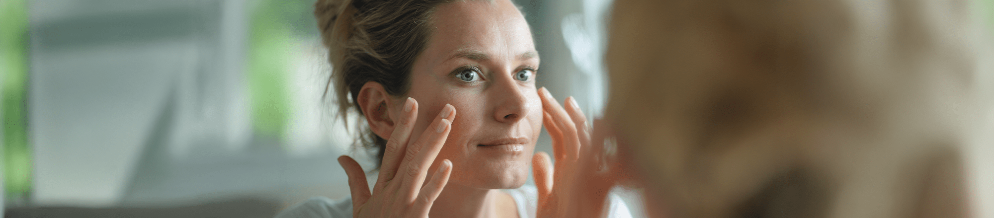 Eine Frau trägt Creme auf ihr Gesicht auf und blickt dabei in den Spiegel.