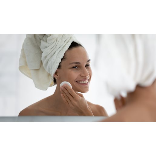 Frau blickt in ihr Spiegelbild und reinigt mit einem Wattepad ihre Wange. Sie lächelt und trägt ein weißes Handtuch in den Haaren.