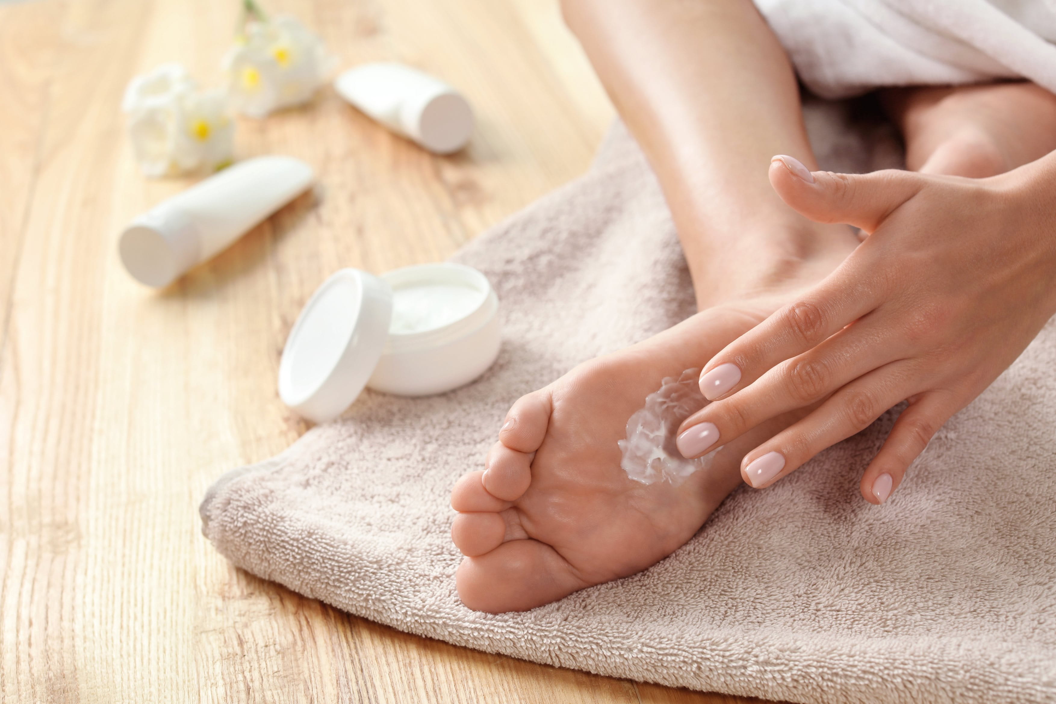 Person sitzt auf einem Handtuch auf dem Boden und cremt sich mit einer Hand die Fußunterseite ein. Im Hintergrund sind offene Creme-Dosen und Salben zu sehen.