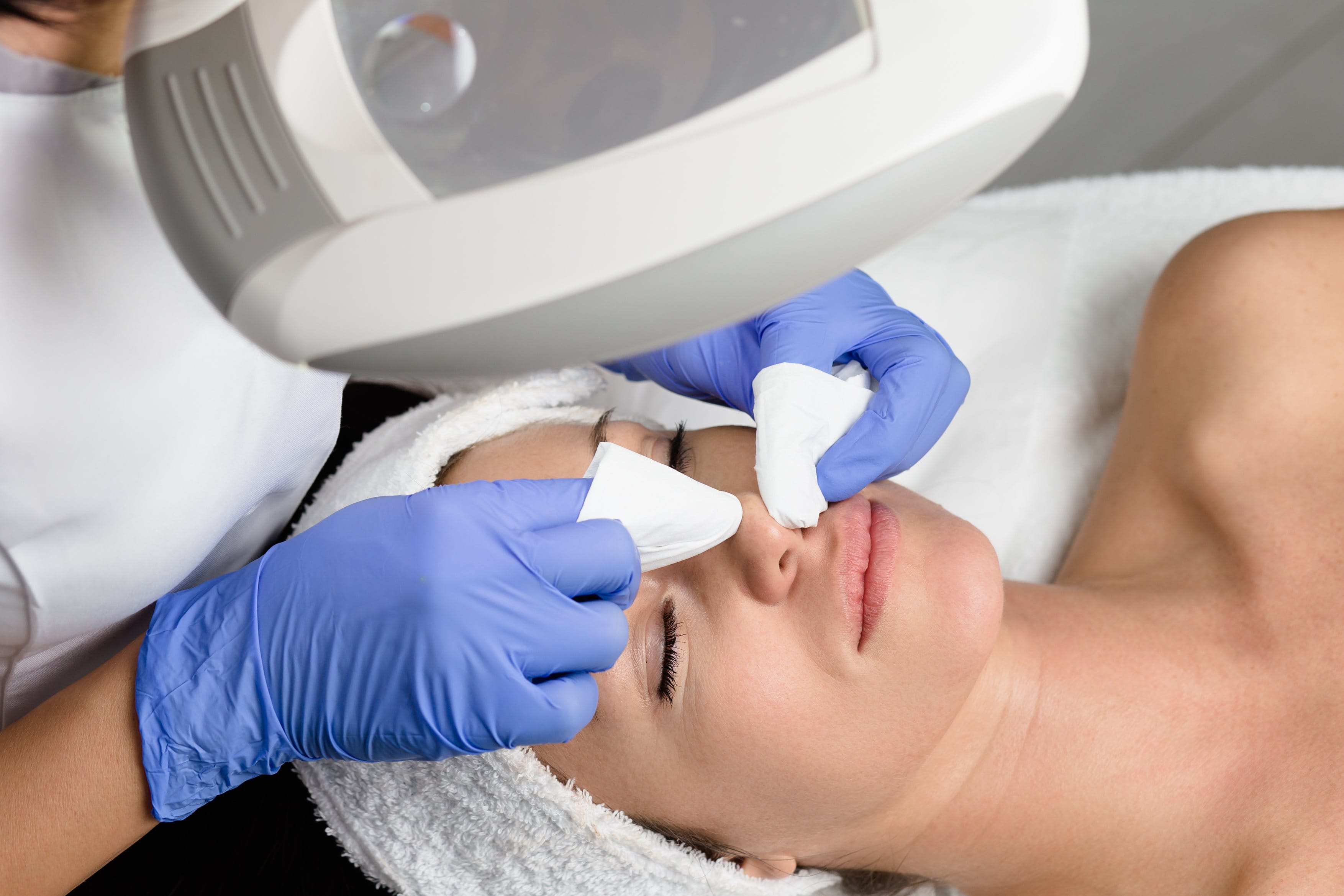 Kosmetikerin trägt Handschuhe und reinigt unter einer Lupenlampe das Gesicht einer Frau, die mit geschlossenen Augen auf einer Behandlungsliege liegt.