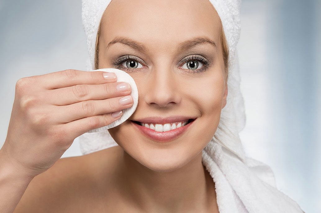 Donna sorride mentre si strucca con un dischetto di cotone, durante la routine di doppia detersione.