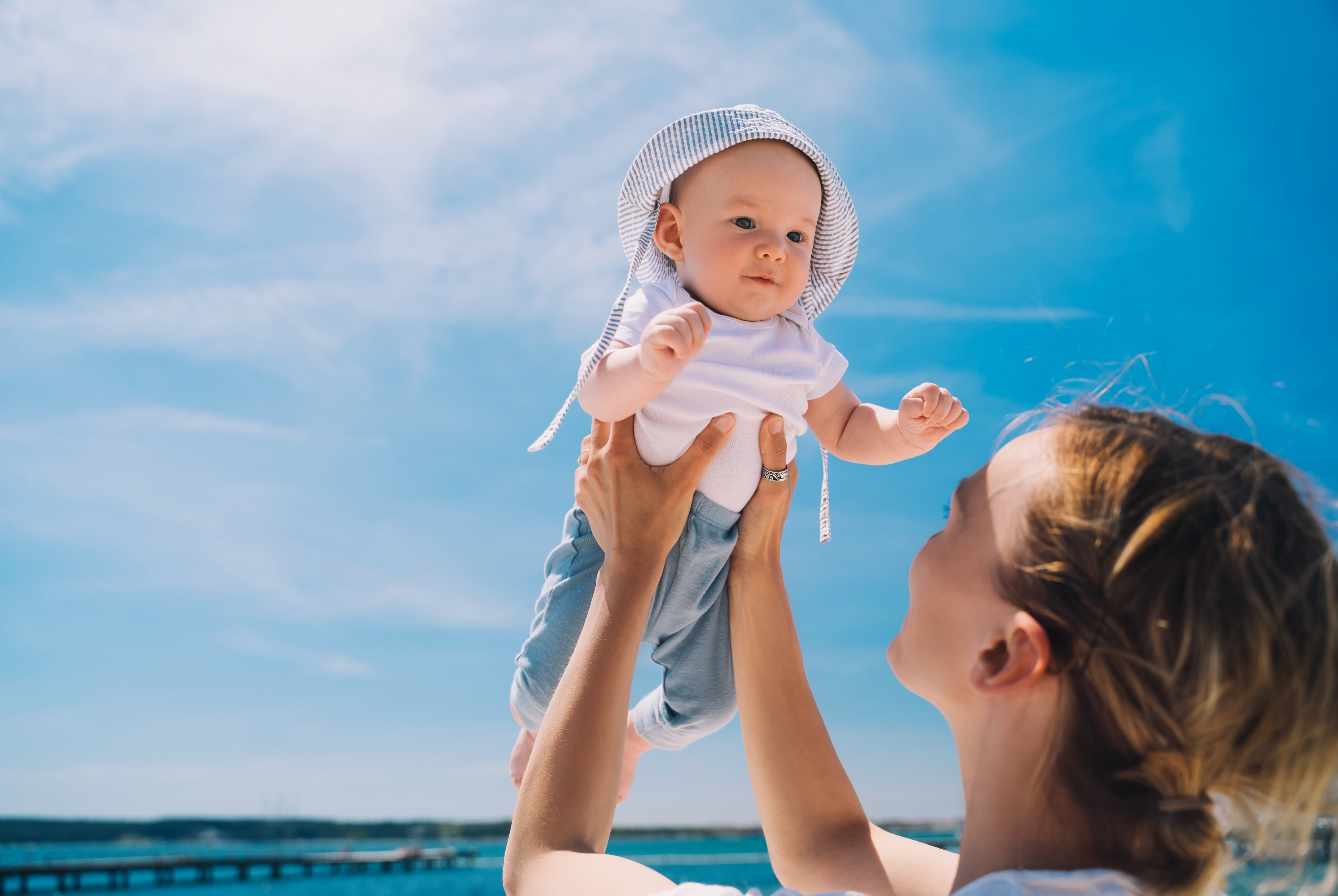 Eine junge Frau hält ihr Baby in die Luft, das einen Sonnenhut und lange Kleidung trägt.