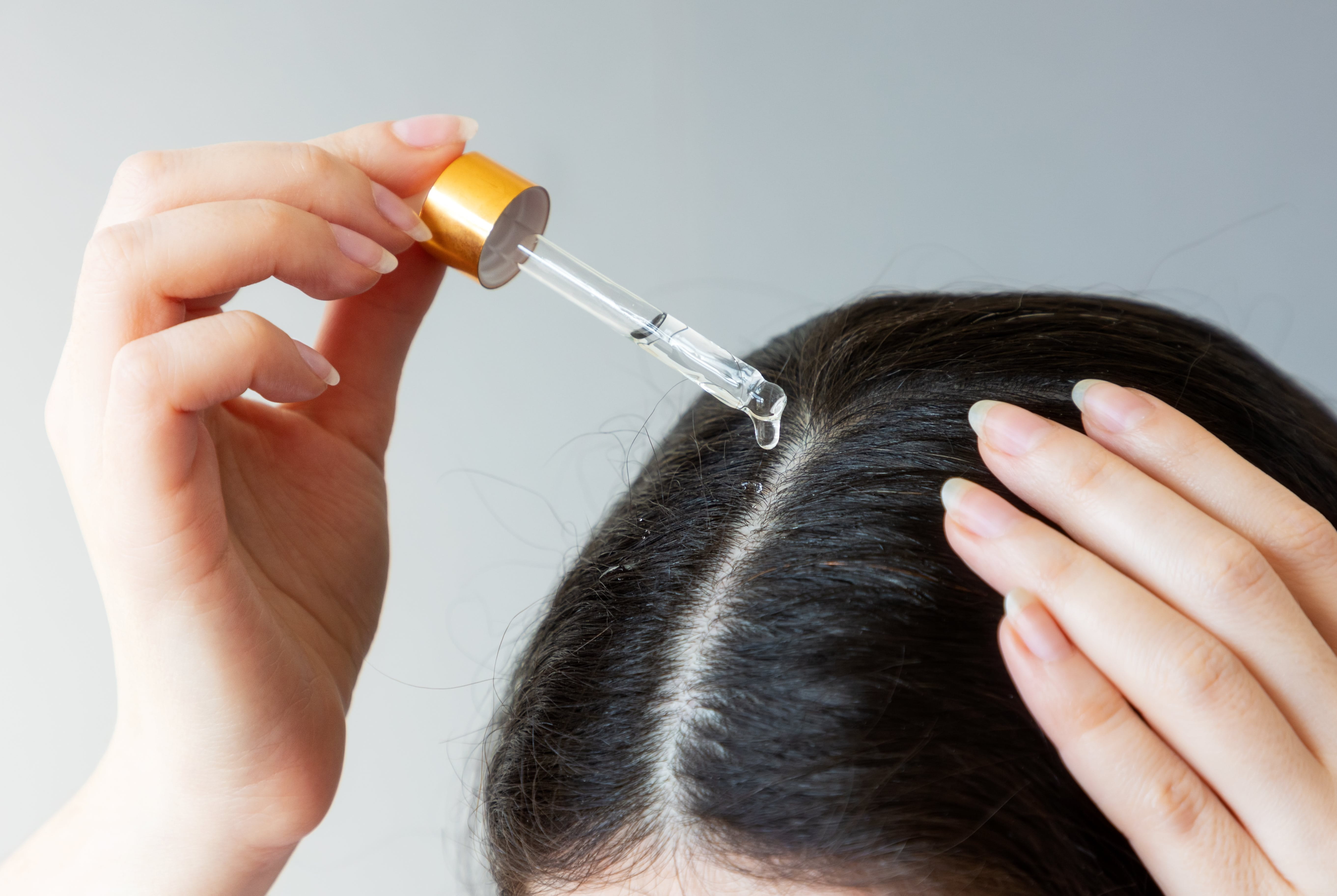 Nahaufnahme des Kopfes einer Frau, die mit der rechten Hand aus einer Pipette Öl auf ihre dunklen Haare aufträgt.
