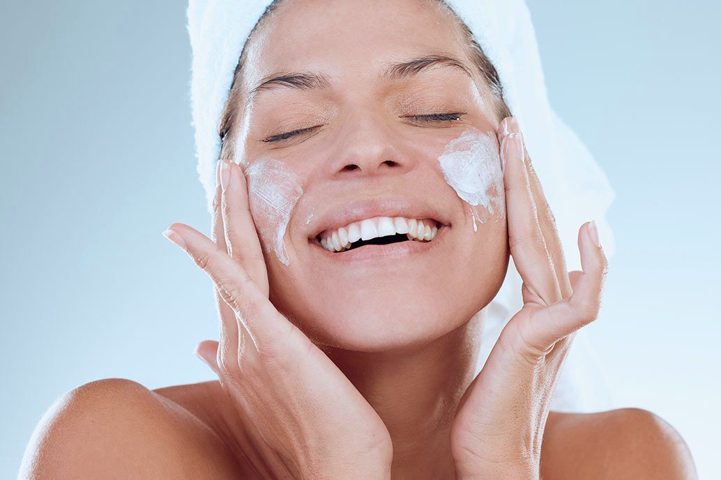 Persona con occhi chiusi massaggia il viso con un detergente schiumoso durante la doppia detersione, mostrando una pelle luminosa e rilassata.
