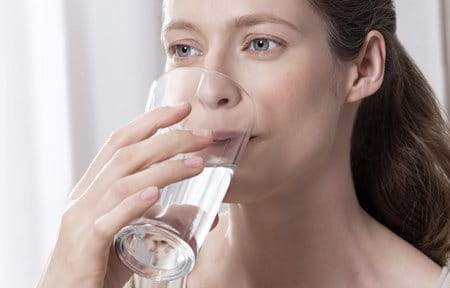 Eine Frau trinkt aus einem Glas mit Wasser, um ihren täglichen Flüssigkeitsbedarf abzudecken. 