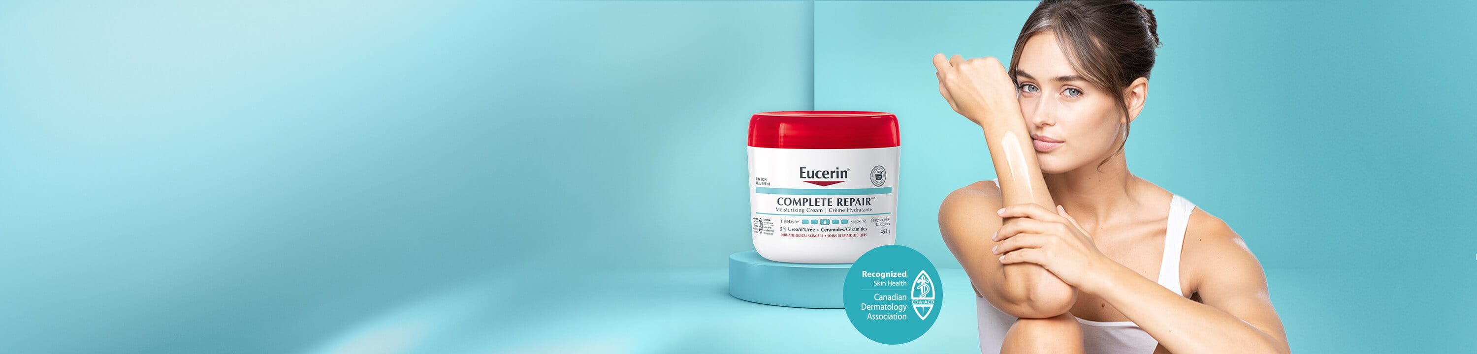 View of a female model wearing a white tank top and sitting down with her knees bent with the Eucerin Complete Repair 454g product in the background.