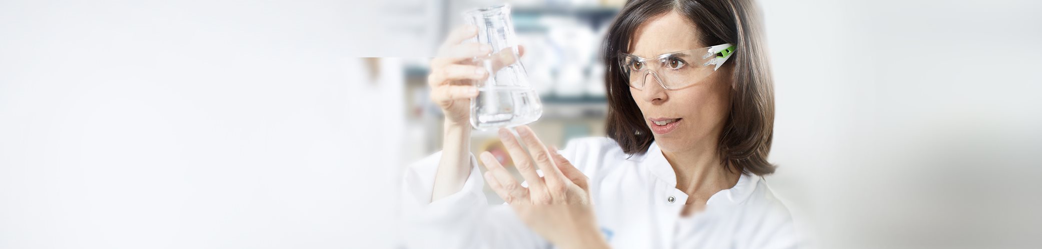 View of a female model wearing protective eyewear and holding up a glass beaker in a lab type setting.