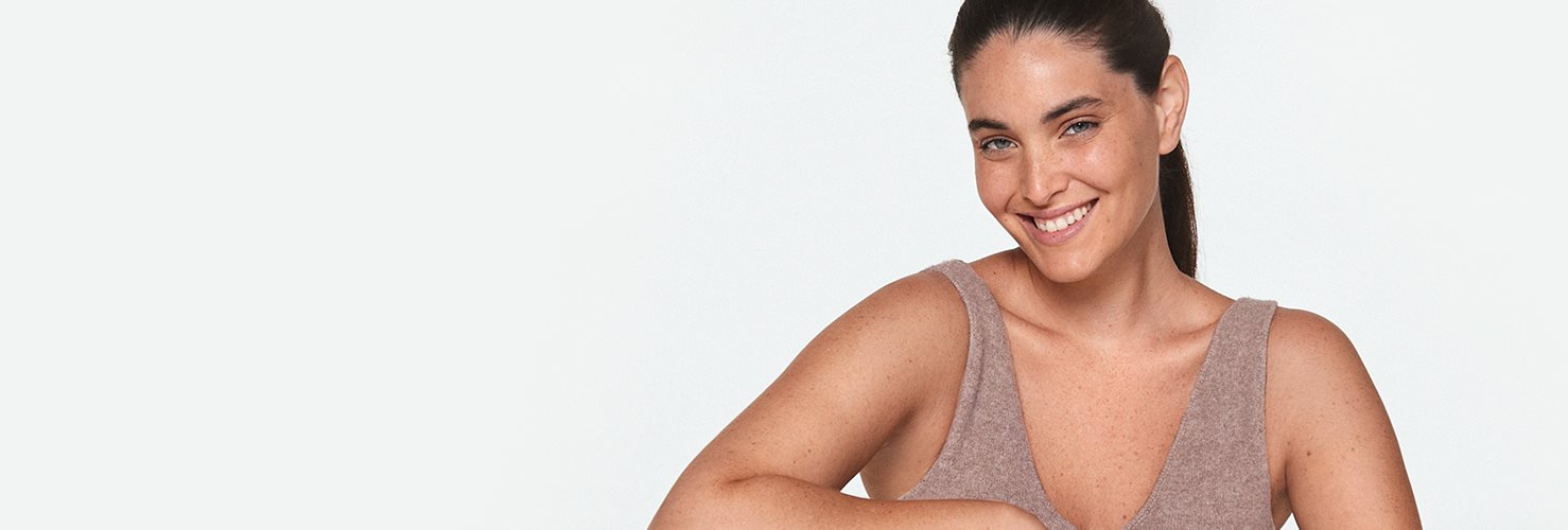 View of a female model with her hair pulled away from her face, wearing a beige coloured tank top while smiling.