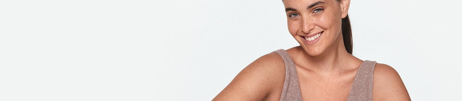 View of a female model with her hair pulled away from her face, wearing a beige coloured tank top while smiling.