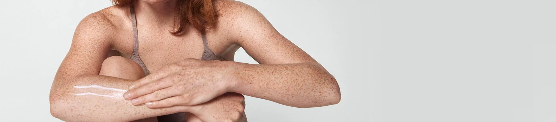 View of a female model with short red hair smearing a cream product along her right arm.