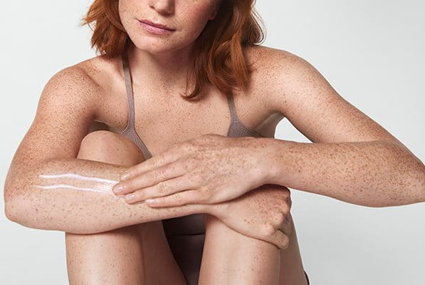 View of a female model with short red hair smearing a cream product along her right arm.