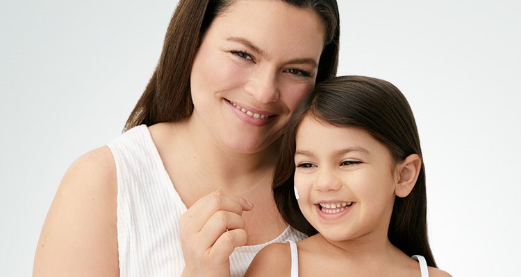 Gros plan d'une femme mannequin et d'une fillette  qui sourient, tandis qu'on voit une touche de  produit en crème sur la joue droite de la fillette.