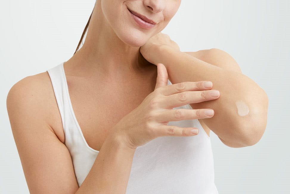View of a female model smiling with a cream product smudged on her left elbow against a grey background.