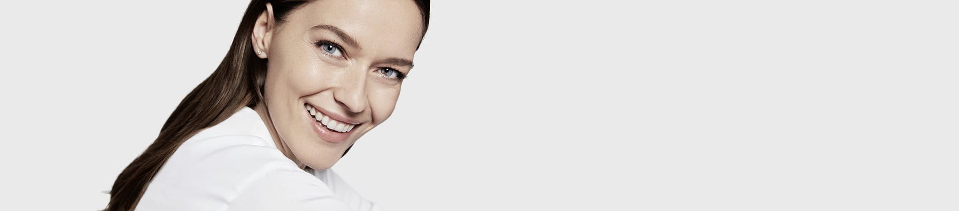 Sur un fond d'écran blanc, une femme mannequin aux cheveux bruns et aux yeux bleus affiche un grand sourire. Elle porte un genre de chemisier blanc.