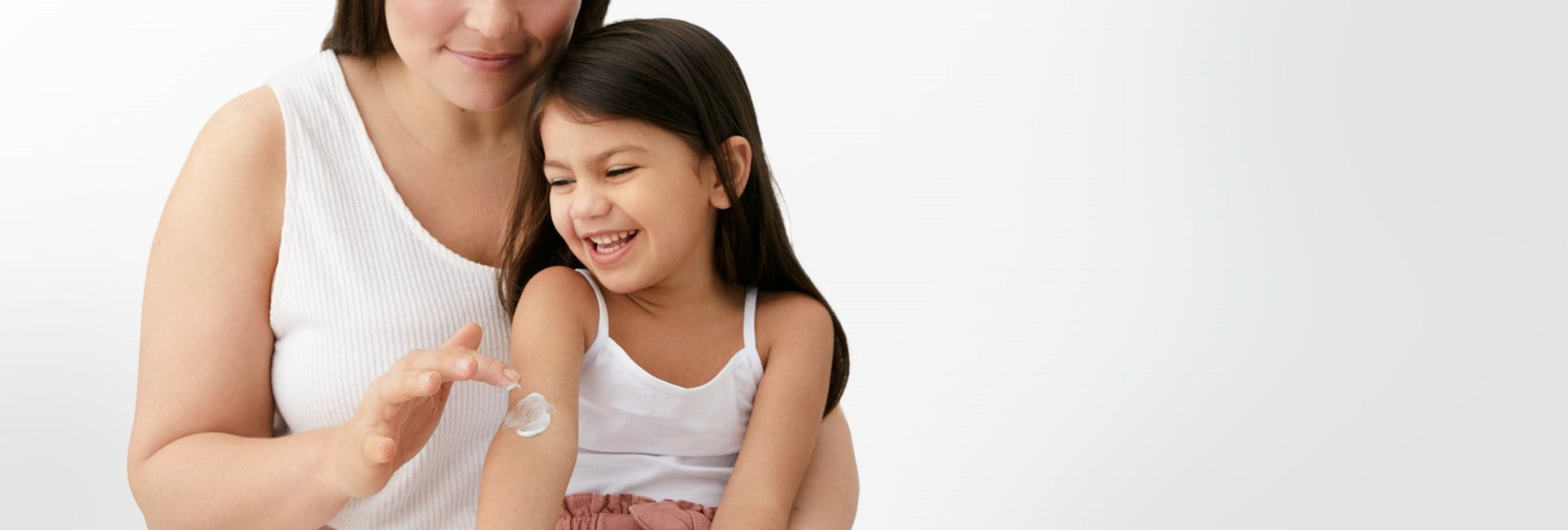 A closeup view of a female model and a little girl smiling with a cream product being applied to her right arm.