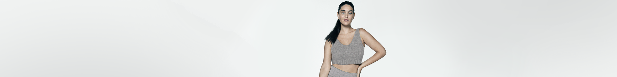 View of a female model with her hair pulled back and wearing a grey coloured knit top with her left hand pressed against her left hip.