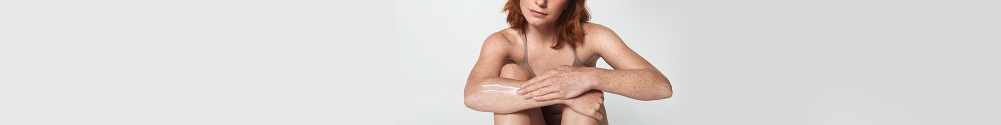 View of a female model with short red hair smearing a cream product along her right arm.