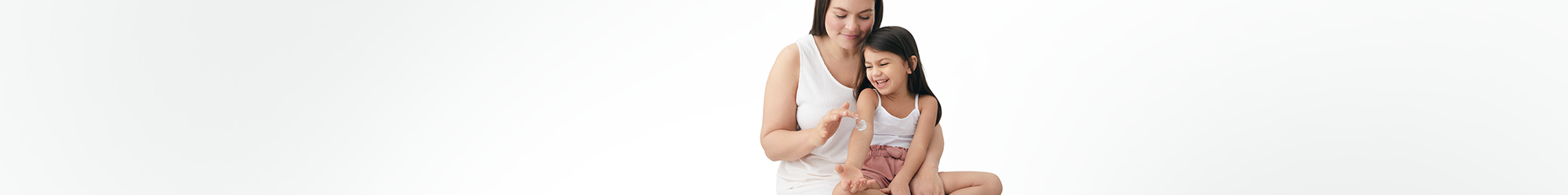 View of a female model applying a cream product on a little girl's arm against a white background.