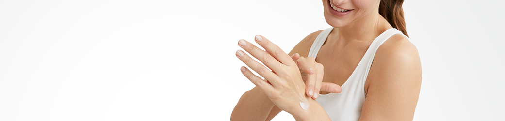 Une femme mannequin portant un débardeur blanc touche une petite quantité de produit en crème sur sa main gauche.