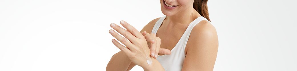 View of a female model wearing a white tank top and applying a cream product to her left hand.