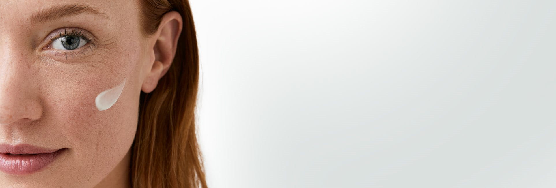 Gros plan du visage d&rsquo;une femme mannequin aux cheveux roux courts, avec sur sa joue gauche une touche de produit en crème.