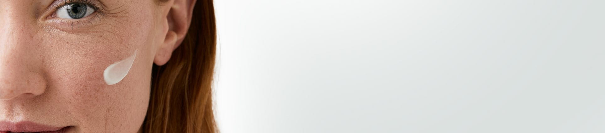 Gros plan du visage d’une femme mannequin aux cheveux roux courts, avec sur sa joue gauche une touche de produit en crème.