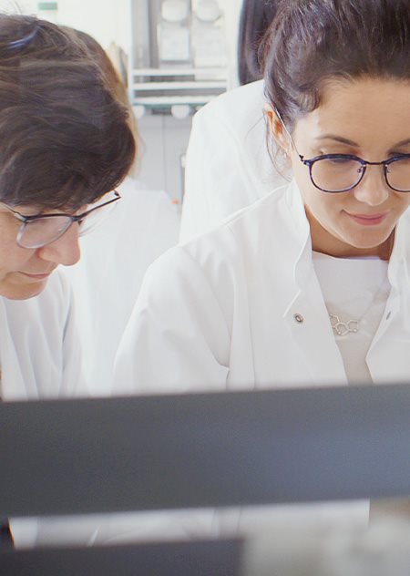 Deux personnes portant des lunettes et une blouse de laboratoire blanche, regardant vers le bas, tandis que d'autres personnes sont montrées derrière elles.