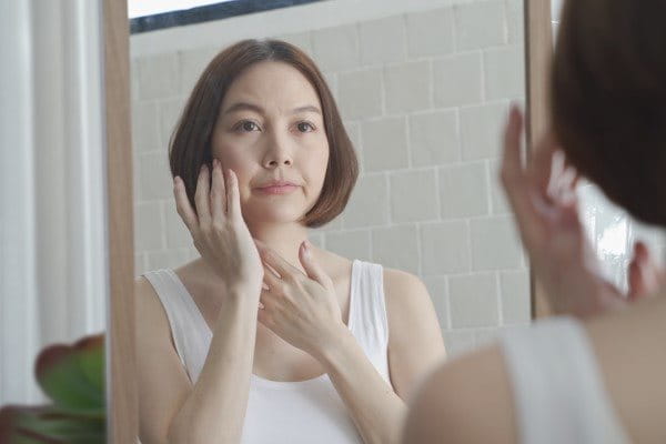 Asiatische Frau mittleren Alters betrachtet ihr Gesicht im Spiegel