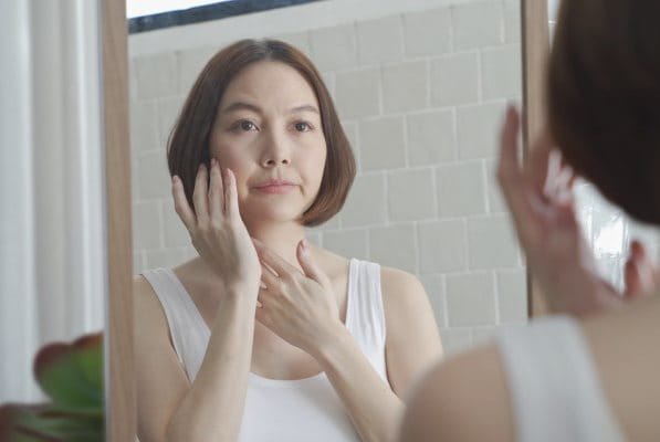 Asiatische Frau mittleren Alters betrachtet ihr Gesicht im Spiegel