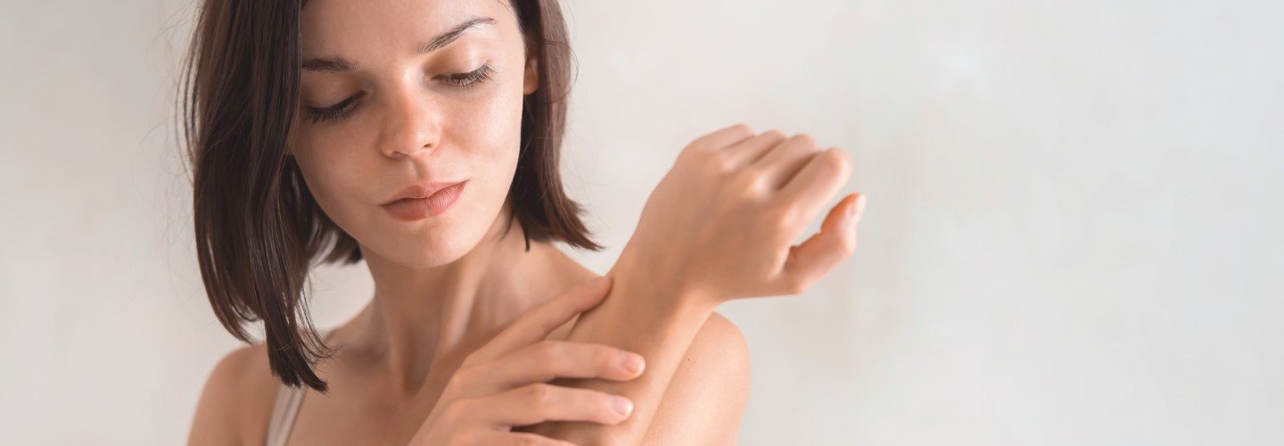 Junge Frau vor hellem Hintergrund, die ihr Handgelenk berührt.