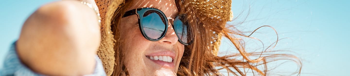 Rothaarige, lächelnde Frau mit Sonnenbrille und Sonnenhut am Strand