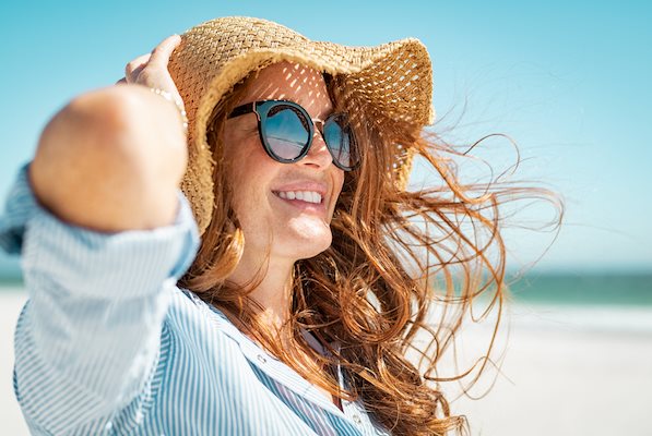 Rothaarige, lächelnde Frau mit Sonnenbrille und Sonnenhut am Strand