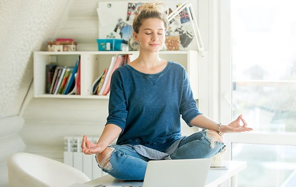 Frau im Home-Office sitzt in Yogahaltung auf dem Schreibtisch