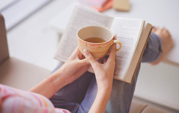 Person holding a cup of tea while reading a book to relax