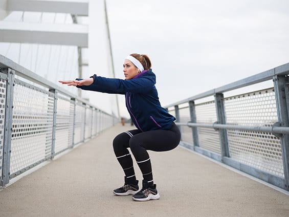 Frau steht in Outdoor-Sportklamotten auf einer Brücke und macht Kniebeugen