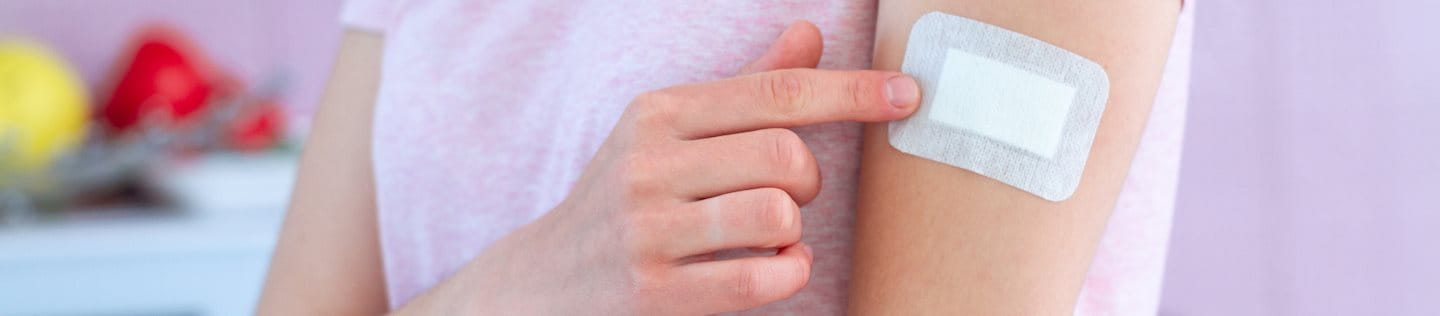 Eine Frau im pinken T-Shirt zeigt auf ihr Pflaster am Arm