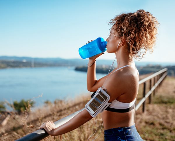 Junge Frau in Sportkleidung steht in der Sonne an einem Geländer und trinkt Wasser