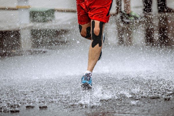 Zu sehen ist der Unterkörper eines Mannes, der durch eine spritzende Pfütze läuft. Er trägt eine kurze rote Trainingshose, blaue Laufschuhe und hat Kinesiologie Tape zur Unterstützung beim Runner's Knee an seine Kniee angebracht. 