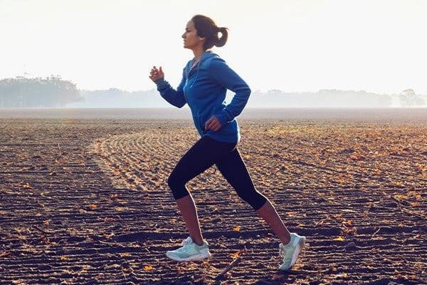 Eine dunkelhaarige weiße Frau läuft mit aufrechter Körperhaltung im Morgenlicht über ein weites Feld. Sie trägt eine blaue Trainingsjacke, Leggins und Laufschuhe, die sie dabei unterstützen kein Läuferknie zu entwickeln. 